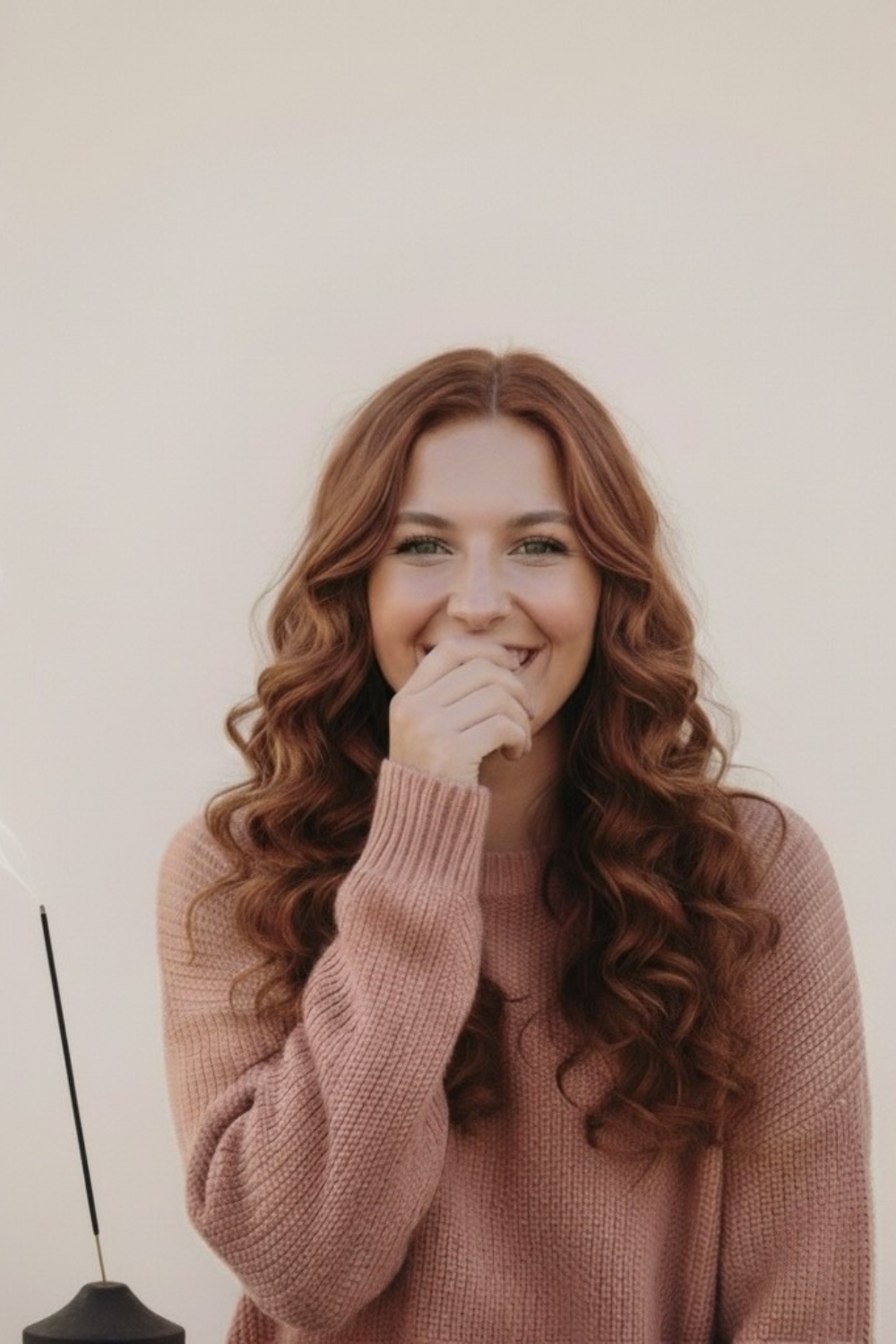 Lachende junge Frau mit geschlossenen Augen hält ein Räucherstäbchen, genießt den Moment der Achtsamkeit und Freude.